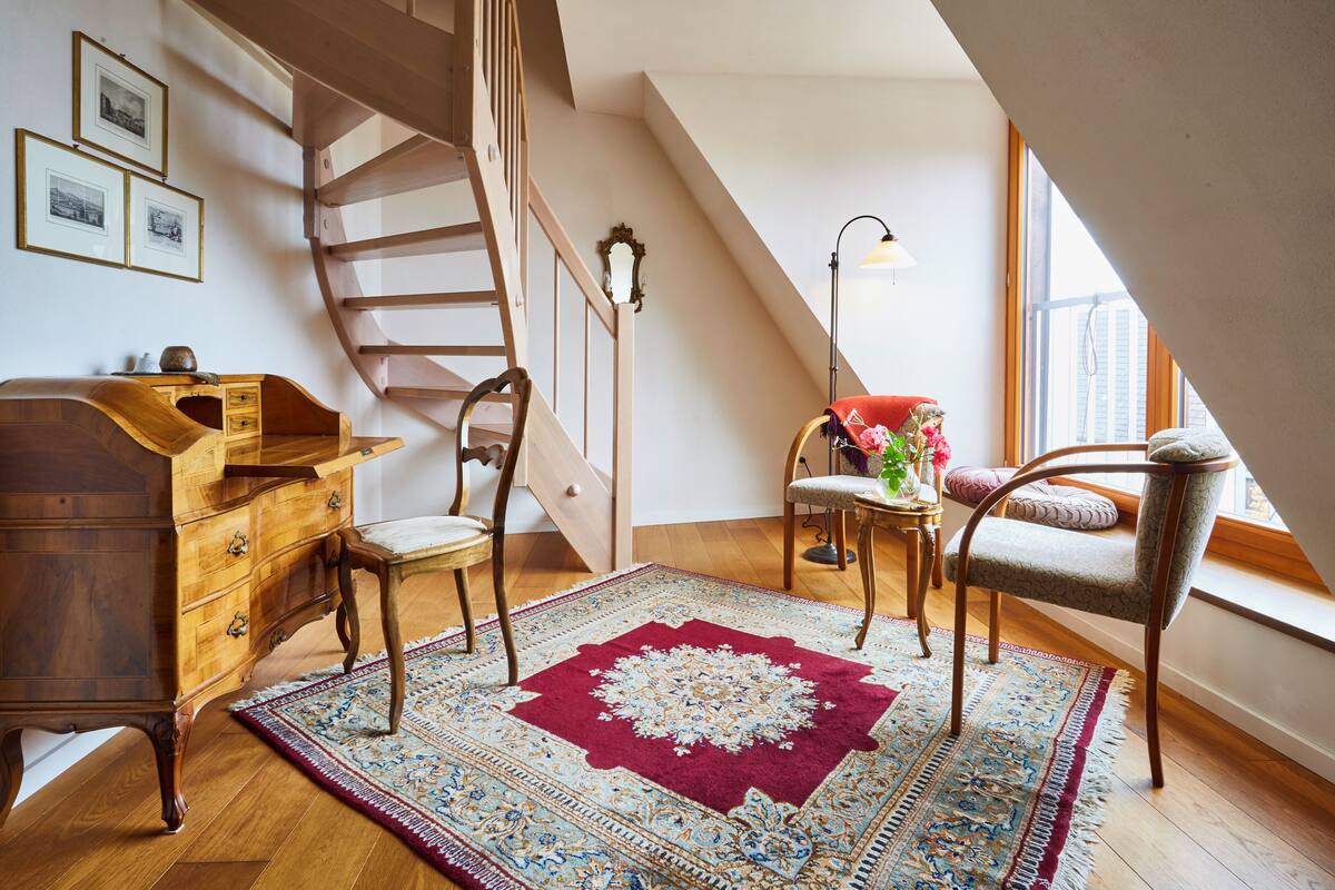 A room with a wooden desk, two chairs, a patterned rug, a staircase, and a window with a view.