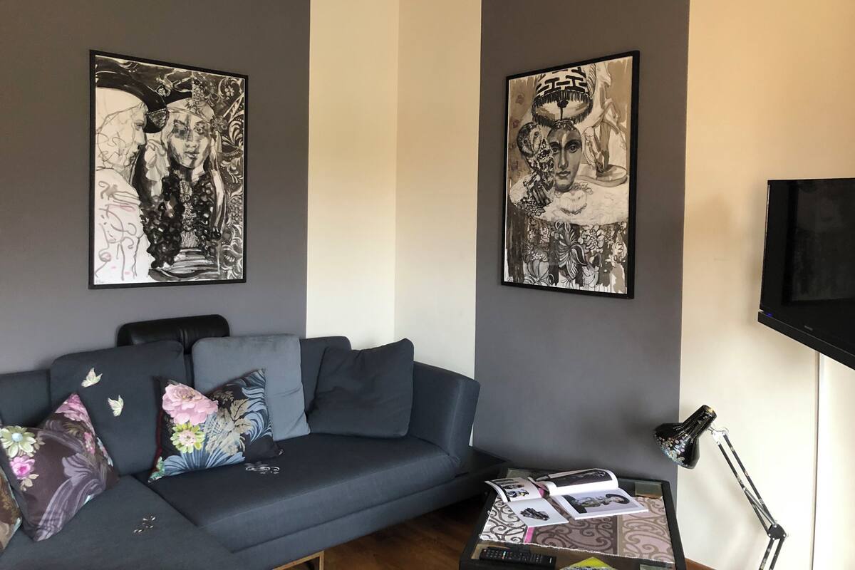 A modern living room with a grey sofa, two large black-and-white artworks on the walls, a flat-screen TV, and a small table with a lamp.