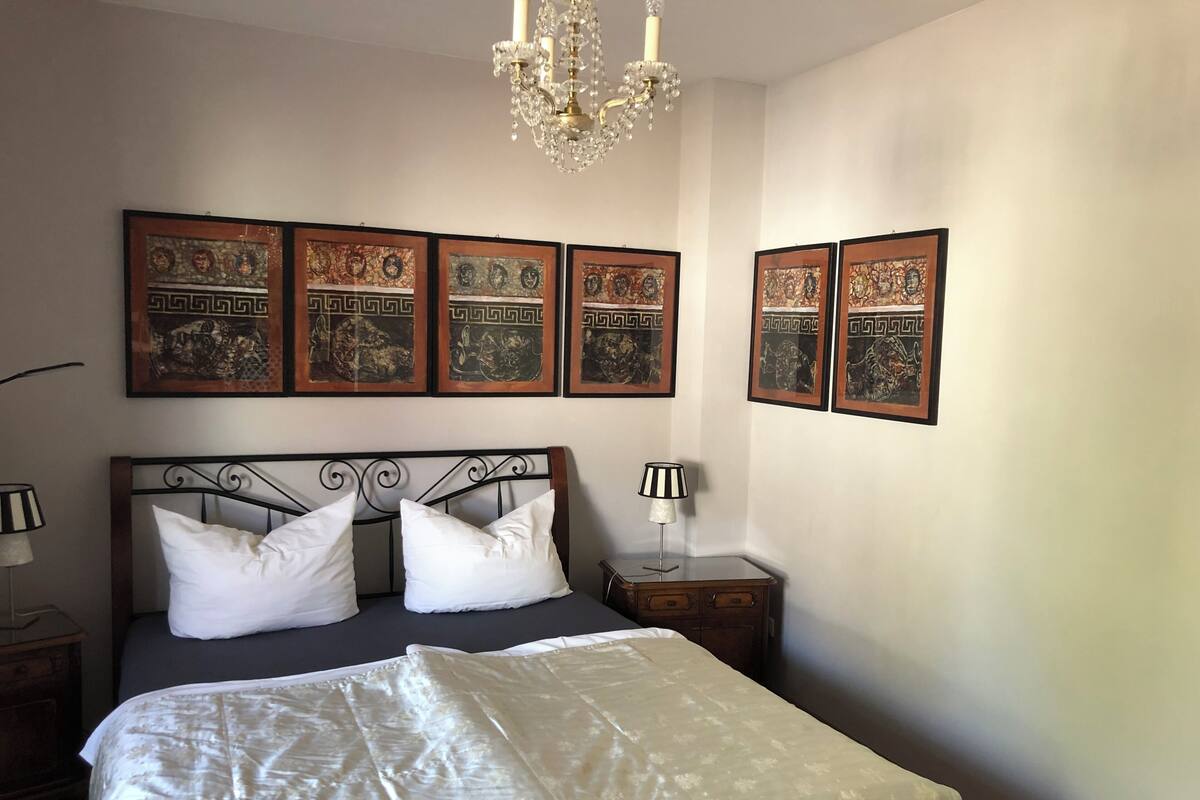 A bedroom with a bed, a chandelier, and framed artwork on the wall.