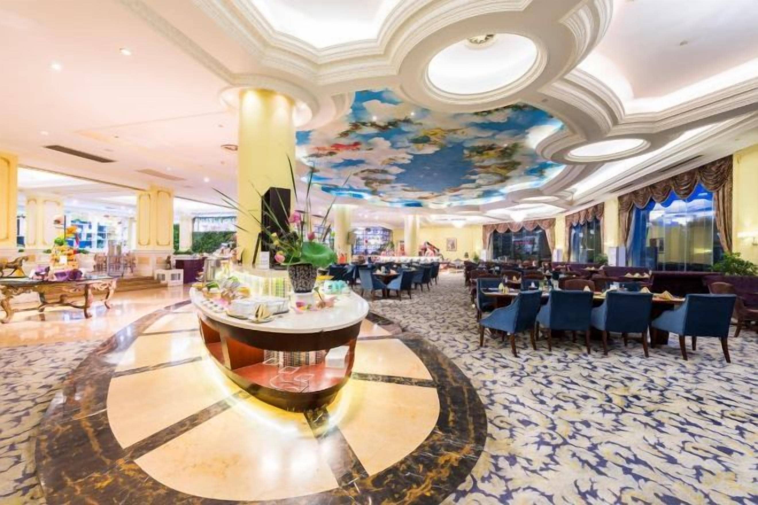 Un vestíbulo de hotel con una mesa redonda central, varias zonas de comedor y un techo con diseño floral.