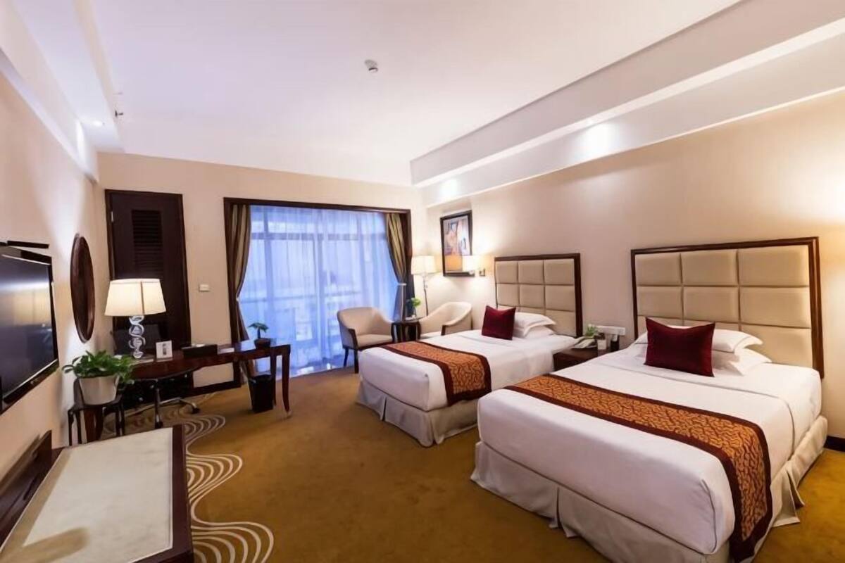 Quarto de hotel com duas camas, uma escrivaninha com cadeira, televisão e janela com cortinas.