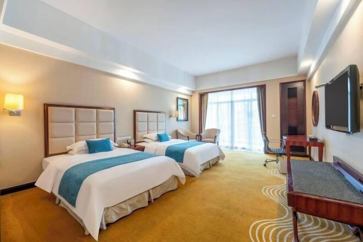 Quarto de hotel com duas camas, uma escrivaninha, uma cadeira, uma televisão e uma janela grande.