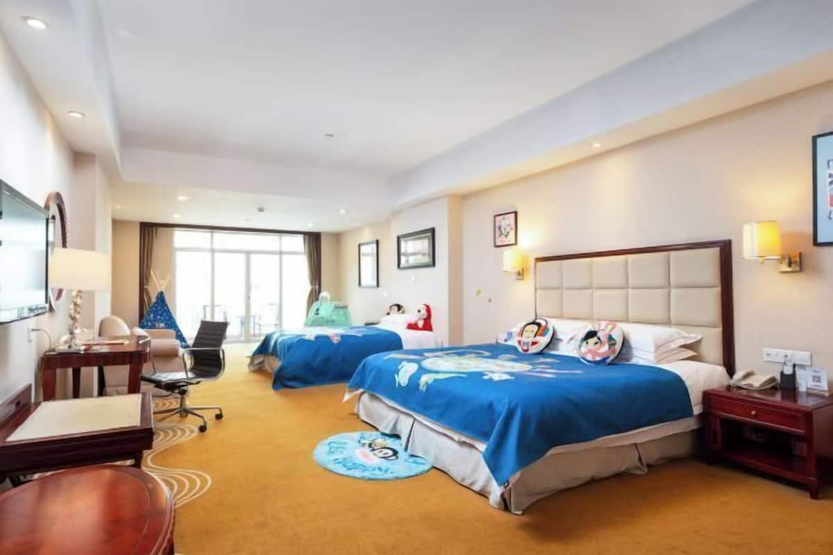 Quarto de hotel com duas camas, uma escrivaninha, uma cadeira, uma mesinha, uma luminária e uma janela.