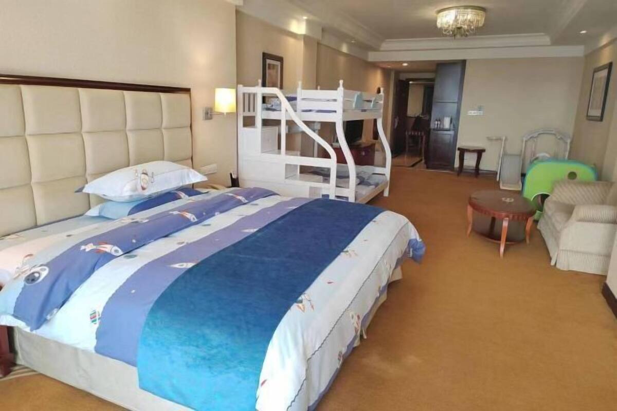 Quarto de hotel com uma cama de solteiro, uma cama separada, uma mesinha, uma cadeira e piso coberto por carpete.