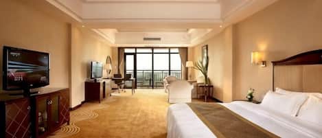 A hotel room with a large bed, a television, a desk with a chair, and a view of the outdoors through large windows.