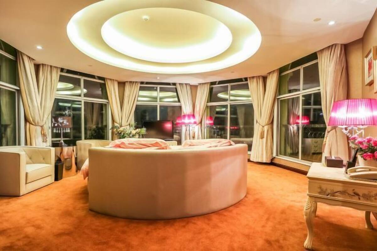 Uma área de estar circular com piso coberto por carpete, grandes janelas com cortinas e um teto com iluminação circular.