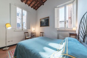 A bedroom with a bed, two windows, a painting, a floor lamp, and a radiator.