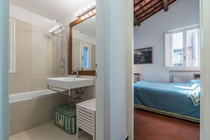 A bathroom with a bathtub, a sink, and a mirror with lights. There is a window with a view of a building outside.