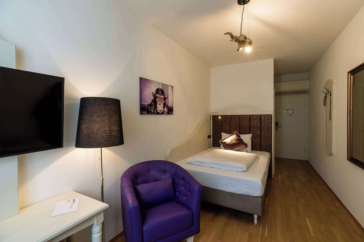 Habitación de hotel pequeña con cama, una silla, escritorio, televisor y un cuadro en la pared.