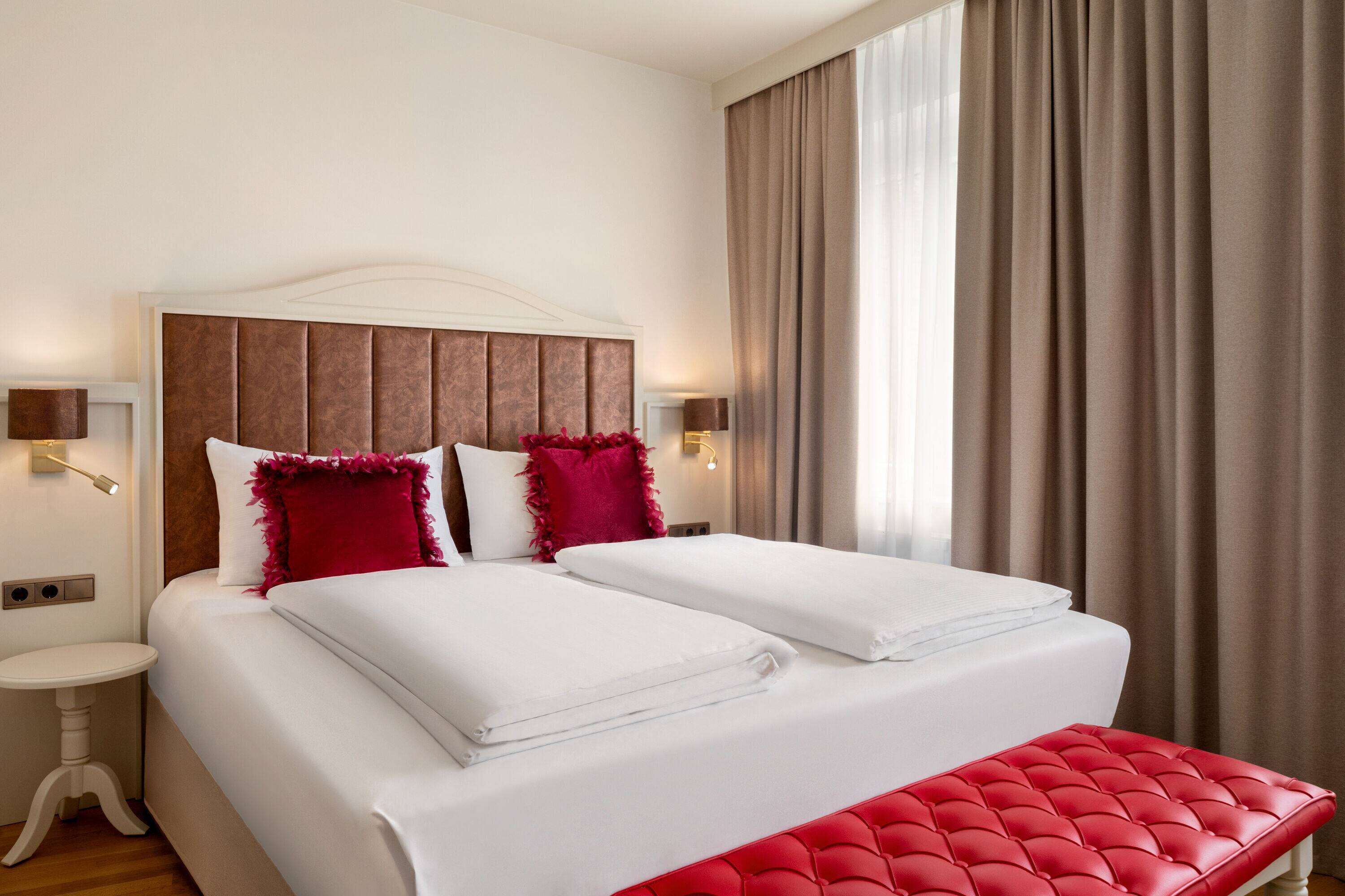 Una cama doble con sábanas blancas y detalles rojos, un banco tapizado en rojo y una mesita de noche con lámpara.