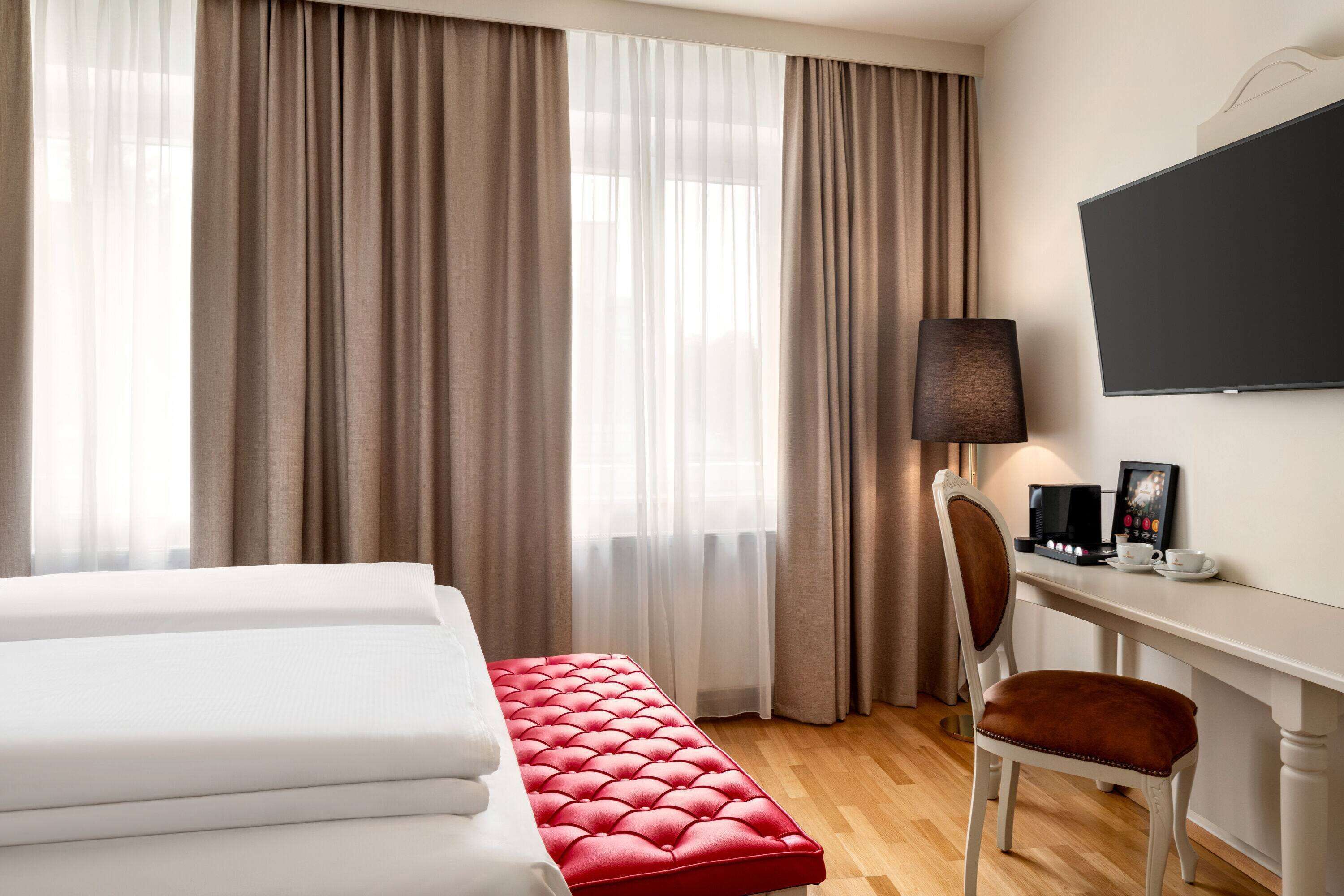A hotel room with a bed, a red upholstered bench, a desk with a lamp, and a flat-screen TV.