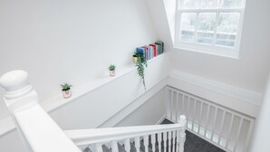 A staircase with a white railing, a small shelf with books and plants, and a window with a view of the outside. - Stunning 2 Bedroom Duplex Apt in Center of Camden (London)
