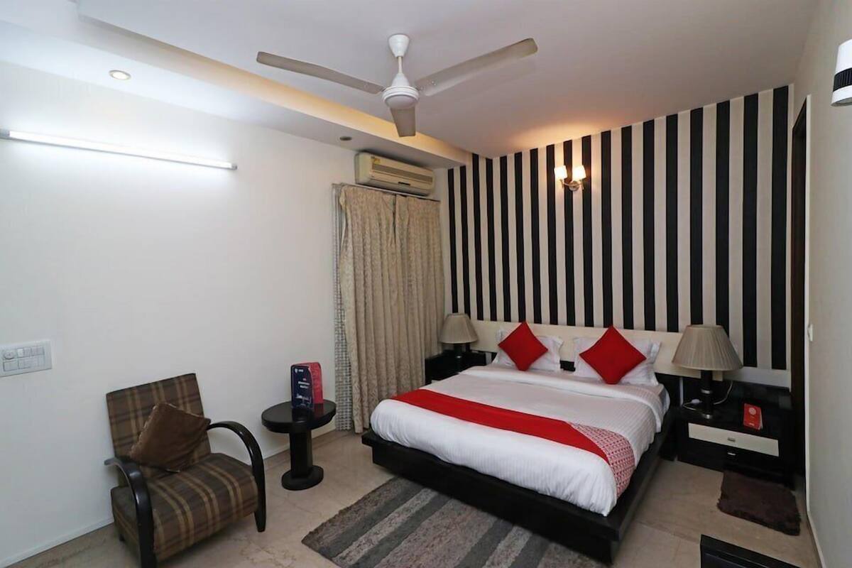 A hotel room with a bed, a chair, a bedside table, a lamp, a ceiling fan, and an air conditioning unit.