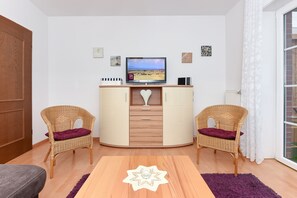 Uma sala de estar com televisão, duas cadeiras de vime, uma mesa de madeira e um radiador branco.