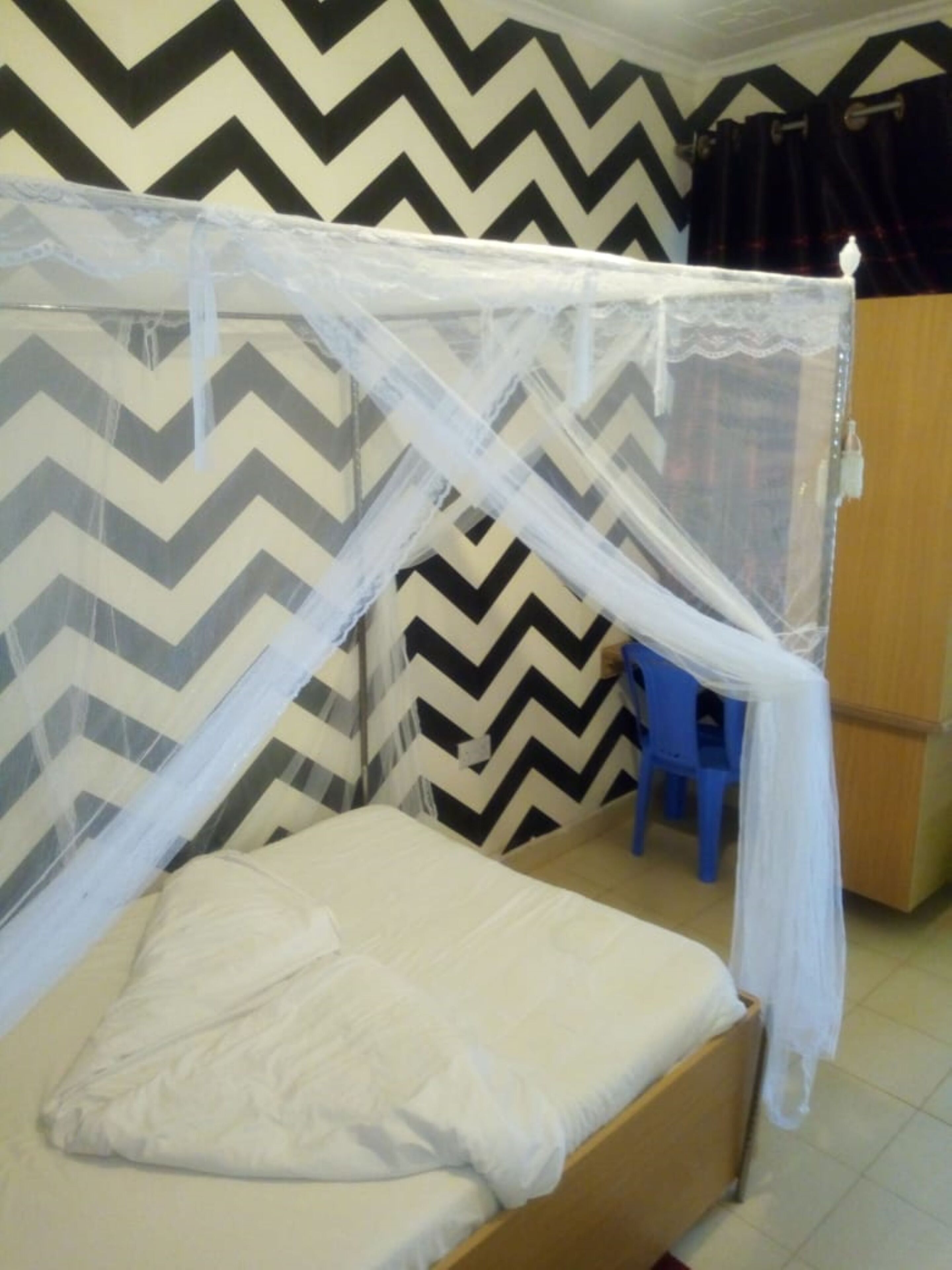 Una cama individual con mosquitero, un taburete azul y un armario en una habitación con paredes de diseño en zigzag.