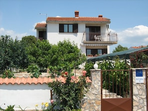 Stunning two-story residence with a balcony and garden area, surrounded by a stone wall and metal gate.