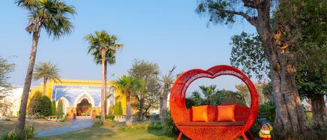 A garden with a red heart-shaped bench and a statue, palm trees, and a building with arches in the background.