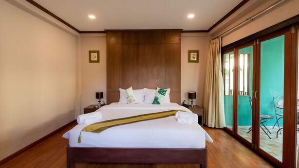 A large bed with a wooden headboard, two bedside tables, and a view of a balcony with a chair. - Vanida Resort - Halal (Ayutthaya)