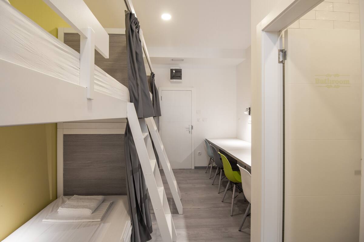 A modern hotel room with a bunk bed, a dining area with chairs, and a bathroom door.