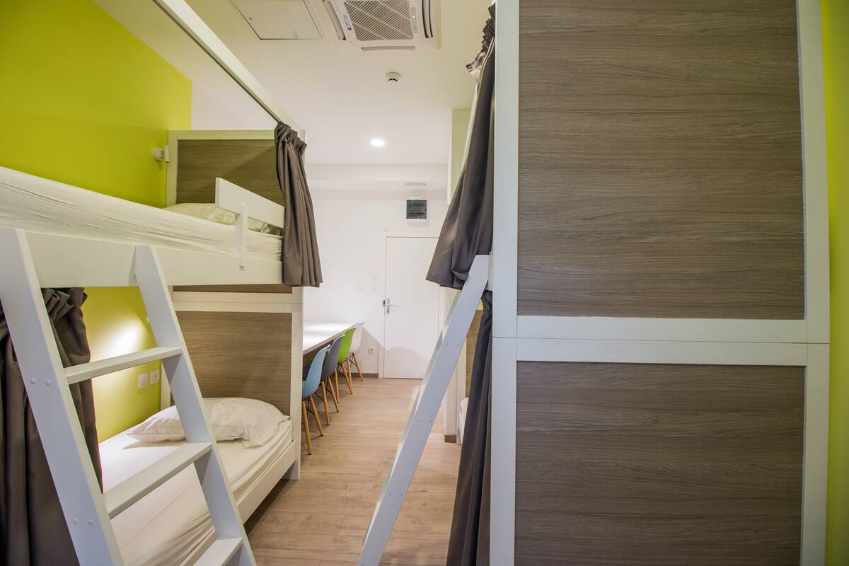 A bunk bed room with a green wall, wooden flooring, and a dining area with chairs and a table.