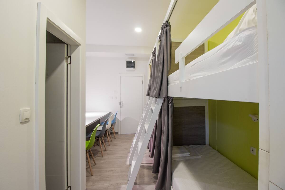 A dormitory room with bunk beds, a desk, chairs, and a door.