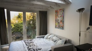Une chambre à coucher avec un lit, un bureau, une lampe sur pied, une fenêtre avec des rideaux et un tableau coloré accroché au mur.