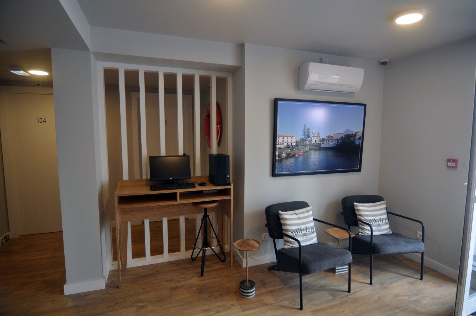 A room with a wooden desk, two chairs, a framed cityscape picture, and an air conditioning unit.