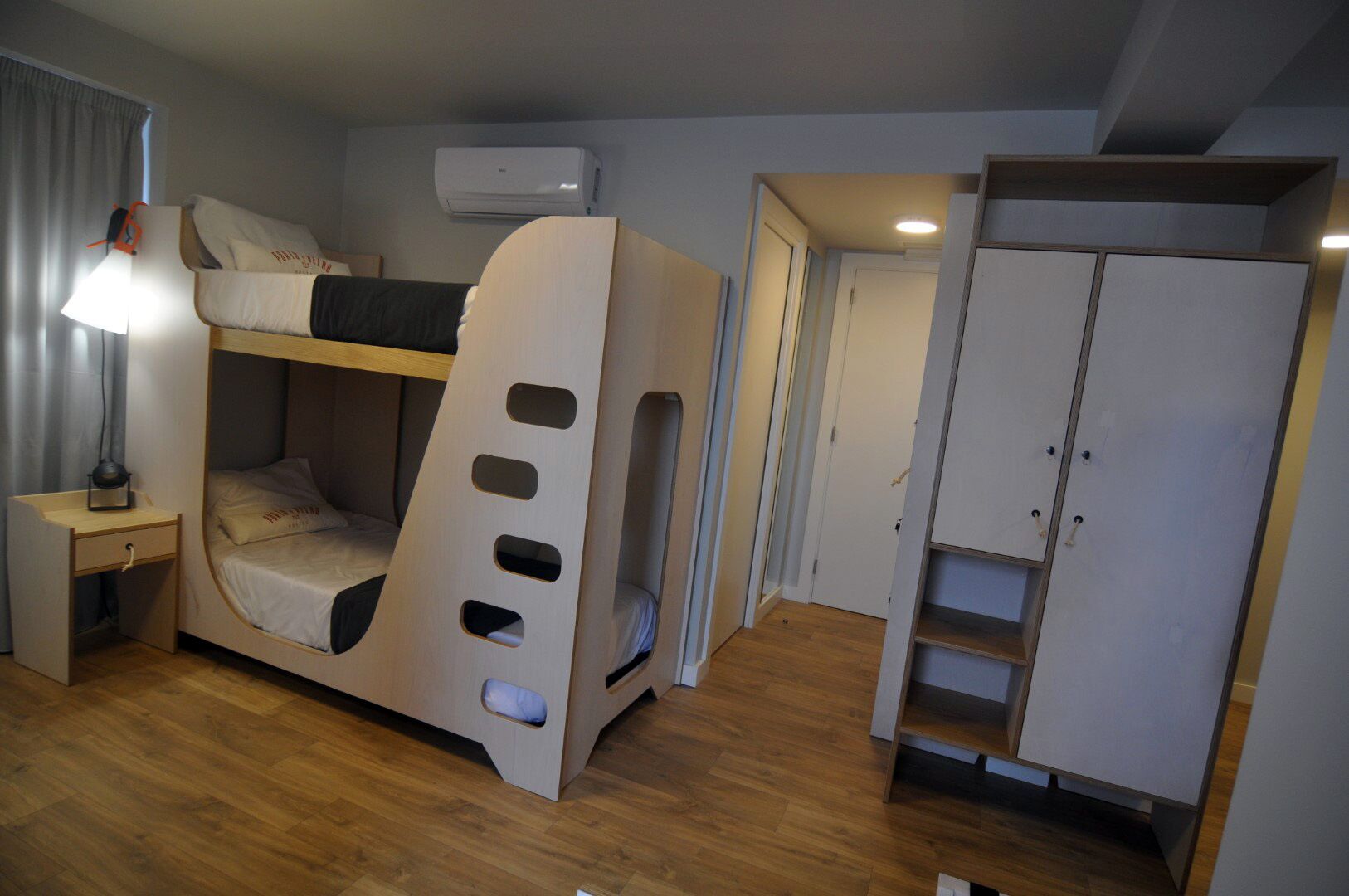 Una cama litera con escritorio y lámpara, un armario y un equipo de aire acondicionado en una habitación con suelo de madera.