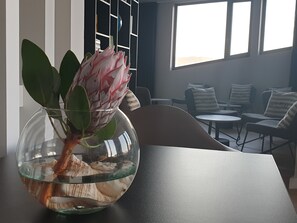Een modern interieur met een glazen schaal met een plant op een tafel, een zithoek met stoelen en een kleine tafel, en een boekenkast op de achtergrond.