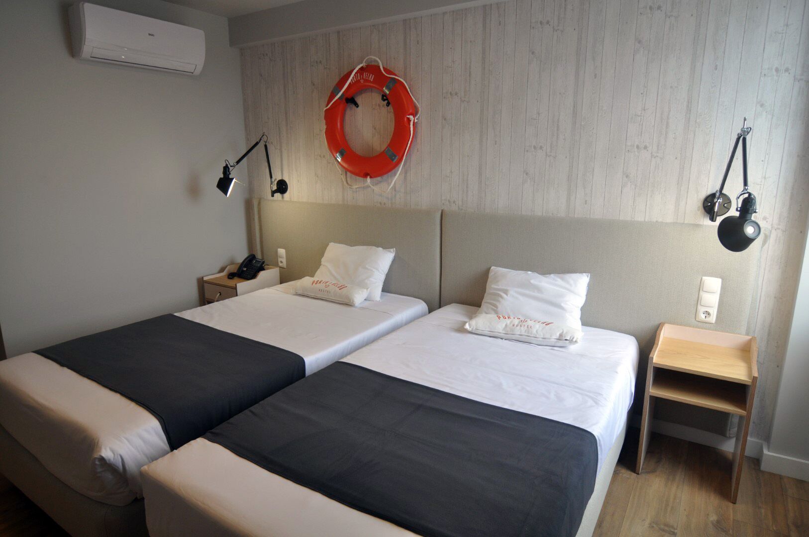 Quarto de hotel com duas camas, cabeceira de madeira, boia pendurada na parede e luminárias de cabeceira.