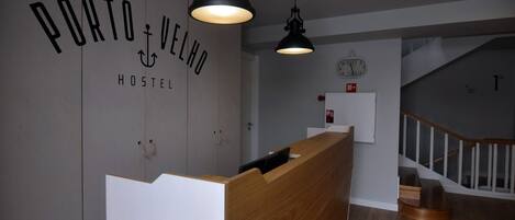 A hostel reception desk with a wooden counter and a black pendant light. A clock is visible on the wall above the counter.