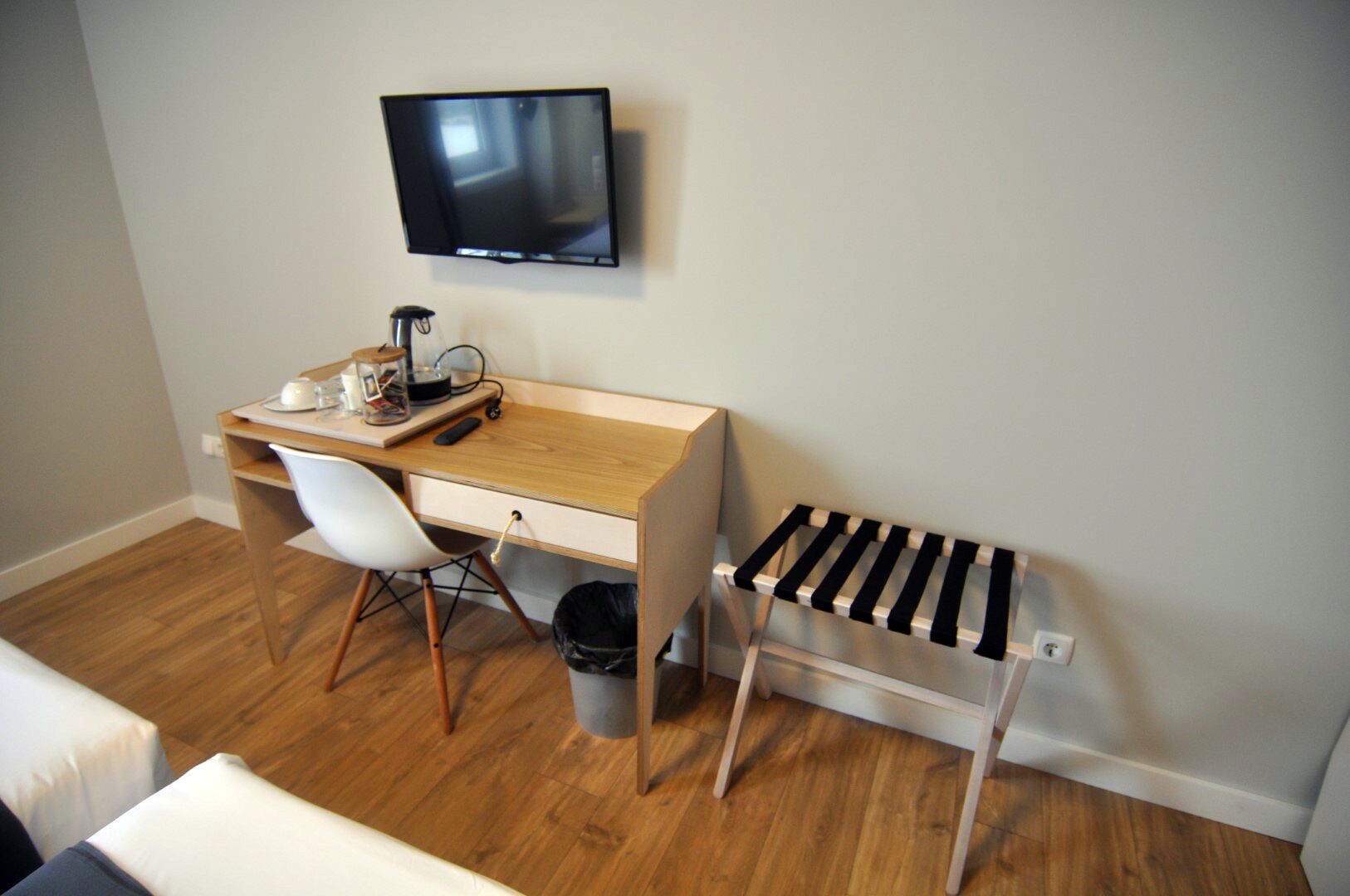 A small, neatly organized room with a wooden desk, a white chair, a television mounted on the wall, and a black and white striped shelf.