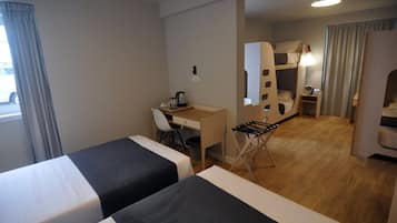 Habitación de hotel con dos camas, un escritorio, una silla, una mesita y una litera al fondo.