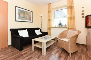 Un salon avec un canapé noir, un fauteuil en osier, une table basse en bois, un lampadaire et une fenêtre avec des rideaux.