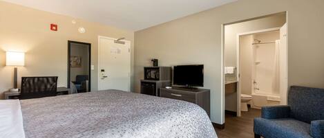 A hotel room with a large bed, a TV, a desk with a lamp, a chair, and a bathroom with a shower and toilet.