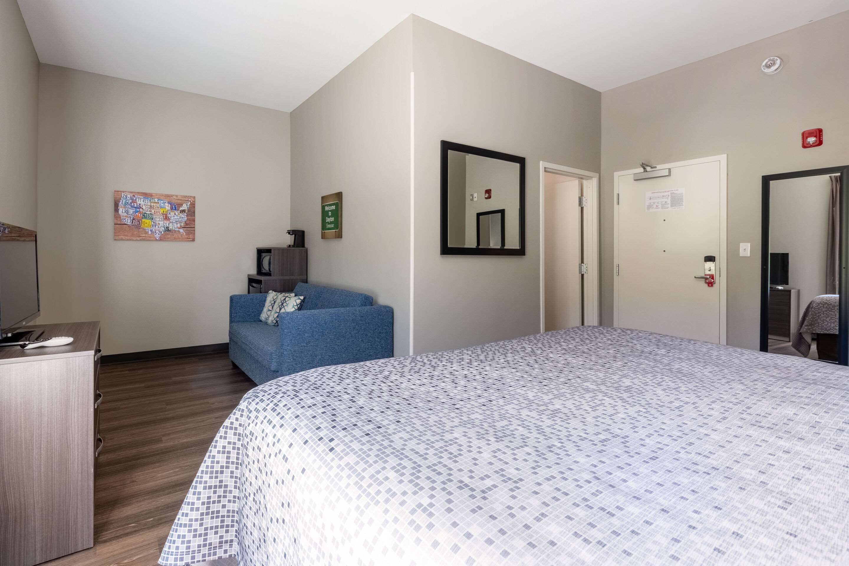 A hotel room with a large bed, a blue sofa, a wooden dresser, a map artwork on the wall, and a door with a sign.