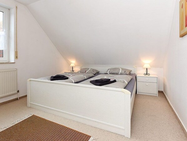 Une chambre à coucher avec un lit blanc, deux tables de chevet avec des lampes, un radiateur et une fenêtre.