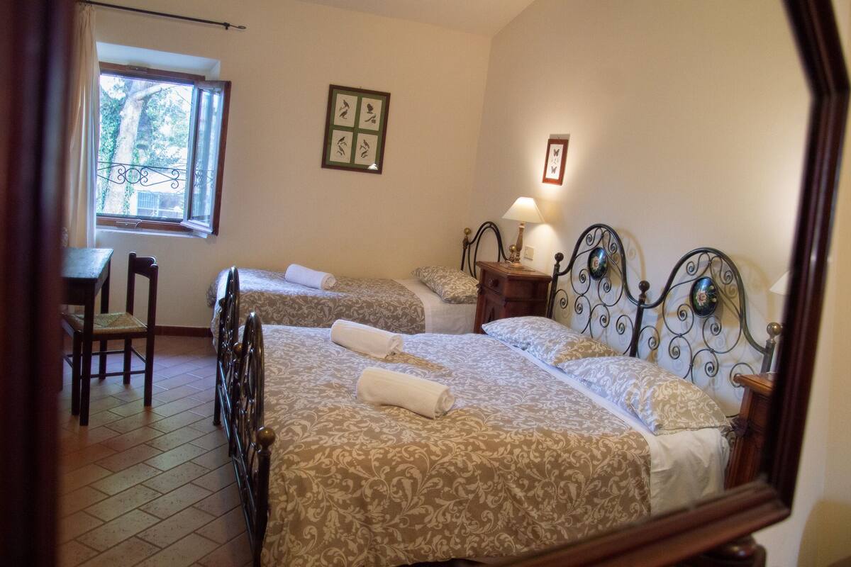 Une chambre d’hôtel avec deux lits, un bureau, une chaise et une fenêtre donnant sur des arbres.