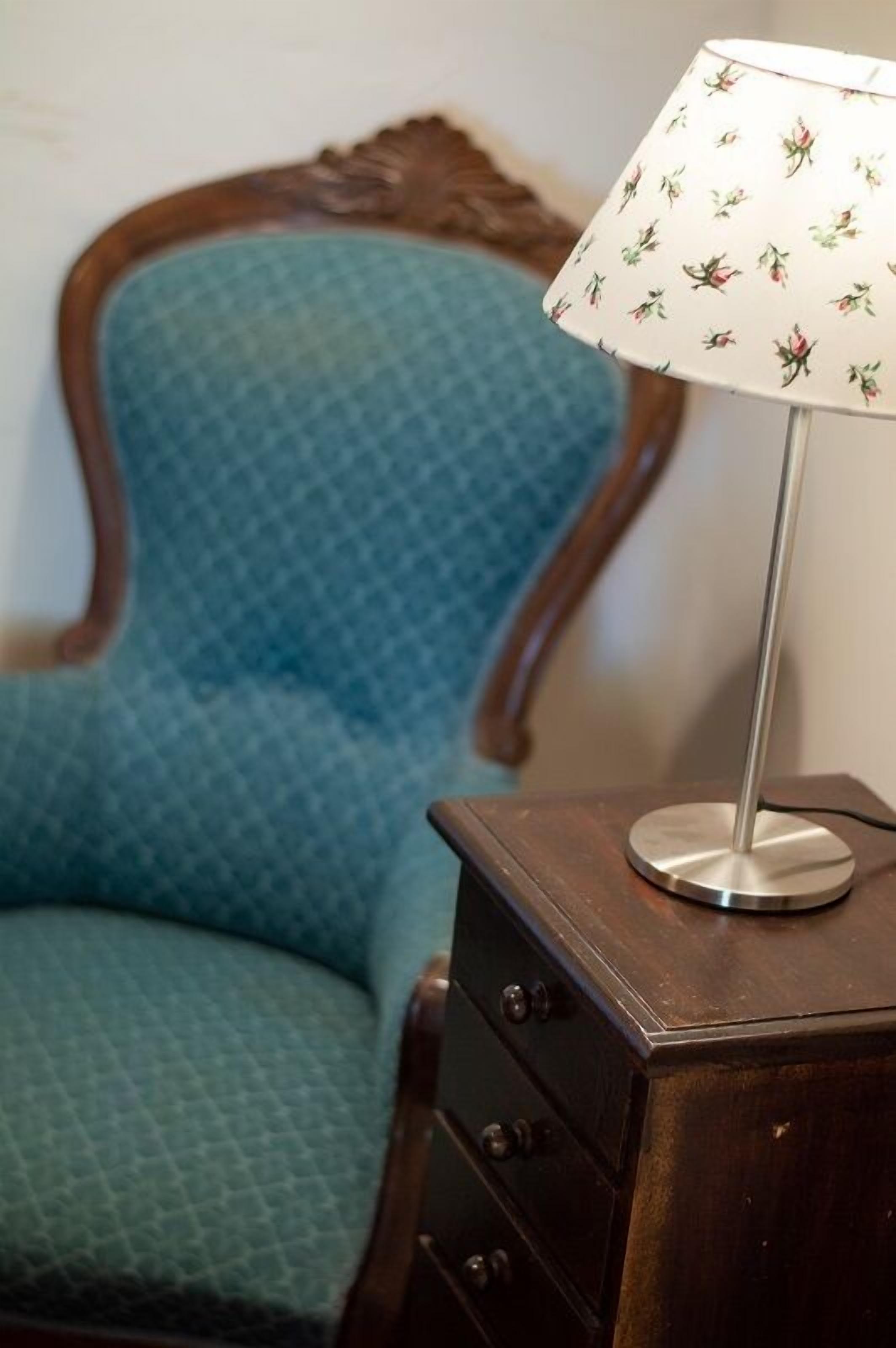 A blue upholstered chair with a wooden armrest and a metal lamp with a floral-patterned lampshade on a wooden nightstand.