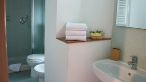 Ein Badezimmer mit Toilette, Waschbecken und Dusche. Handtücher liegen auf einem Regal und ein Korb enthält Pflegeprodukte.