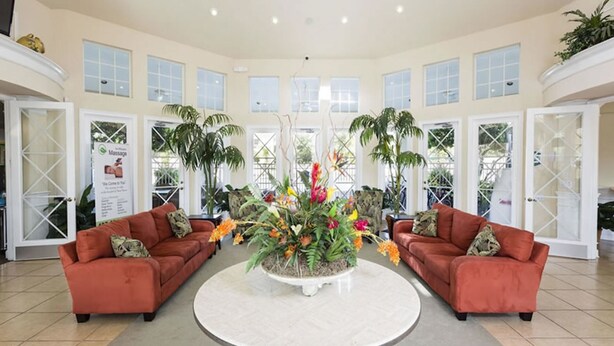 A spacious lobby with two red sofas facing a central round table surrounded by a large floral arrangement. The room has large windows and is decorated with potted plants.