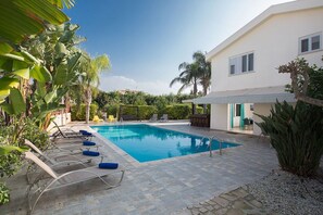 Piscina com espreguiçadeiras, cercada por vegetação e um edifício com fachada branca.
