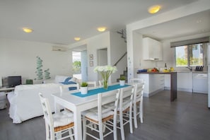 Uma sala de estar moderna e bem iluminada, com uma mesa de jantar, móveis brancos e uma cozinha ao fundo.