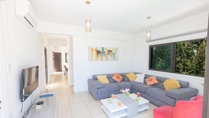 A modern living room with a grey sofa, a pink chair, and a coffee table. There is a TV on a stand, a painting on the wall, and a window with a view of greenery. - Cyprus Villa Near the Beach, Protaras Villa 1244 (Paralimni)