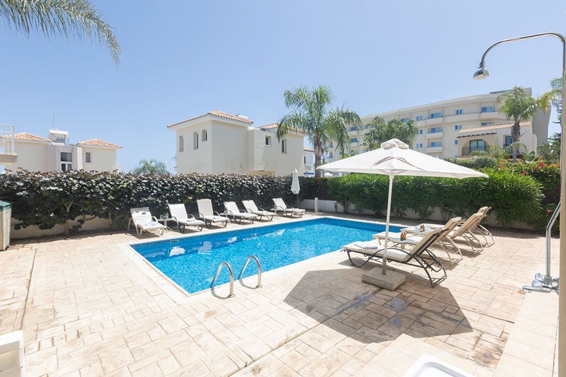 A swimming pool with lounge chairs and umbrellas, surrounded by a hedge and buildings in the background.