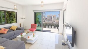A modern living room with a grey sofa, a red chair, and a coffee table. There is a large window with a view of buildings and greenery. - Cyprus Villa Near the Beach, Protaras Villa 1244 (Paralimni)