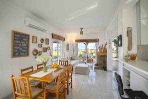 Uma área de refeições ampla com uma mesa e cadeiras de madeira, vista pela porta de vidro, e uma cozinha visível ao fundo.