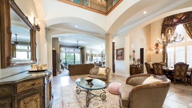 A spacious living room with a high ceiling, a grand staircase, and a dining area with elegant furniture.