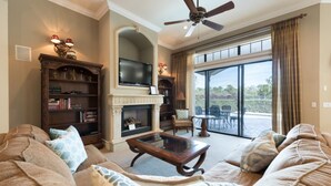 A spacious living room with a fireplace, a ceiling fan, and a large window offering a view of the outdoors.