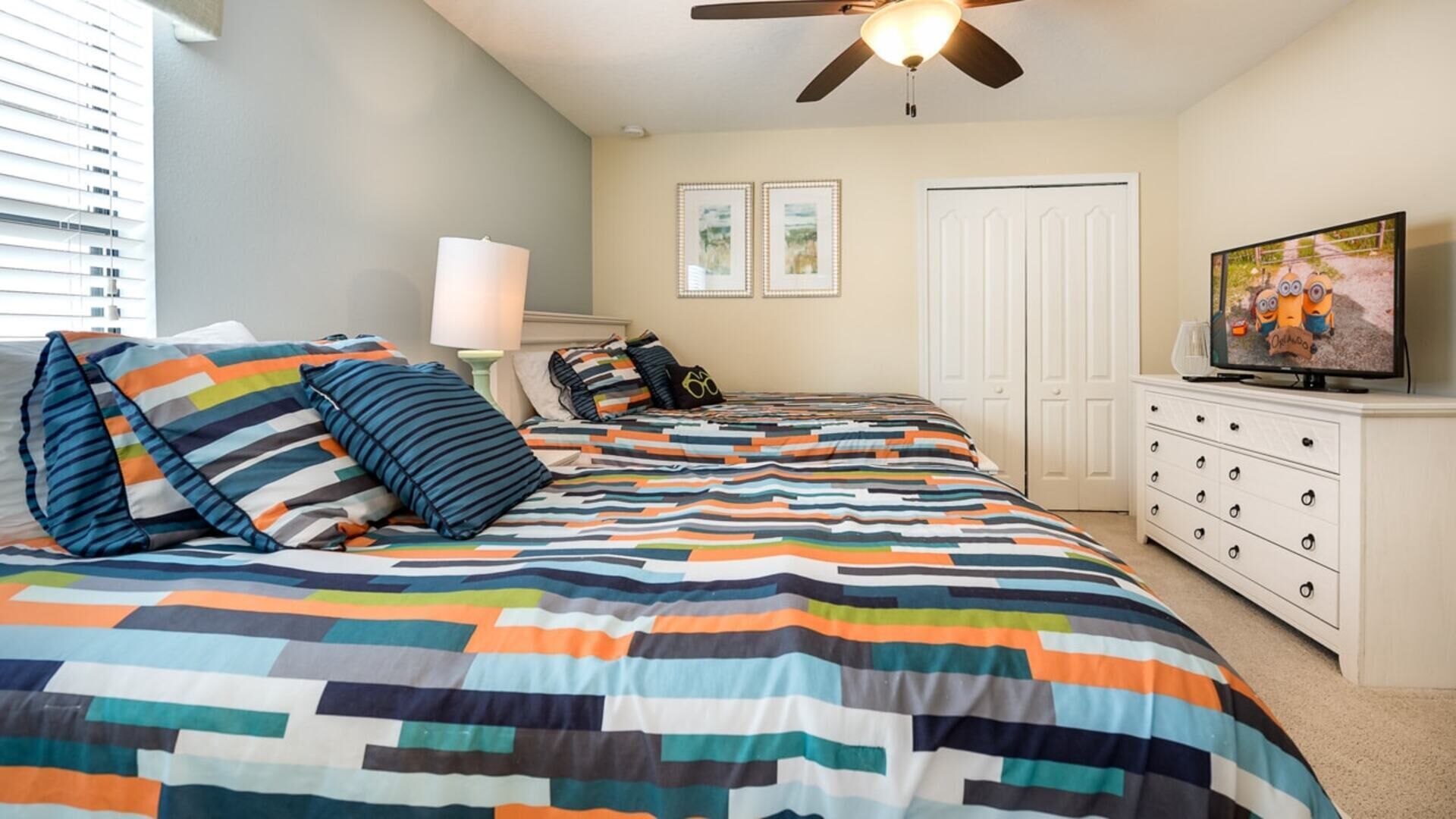 A bedroom with a large bed, a ceiling fan, a TV, a dresser, and a door.